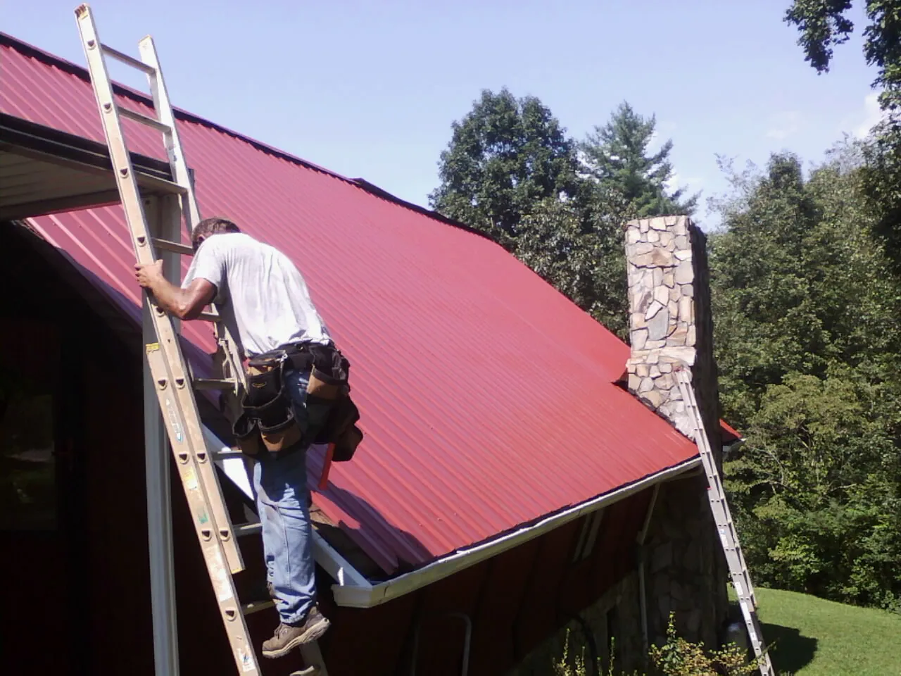 Roofer on a ladder beside a red metal roof and stone chimney — Soffit & Fascia Repair by Roofing Brothers in Sharon Grove